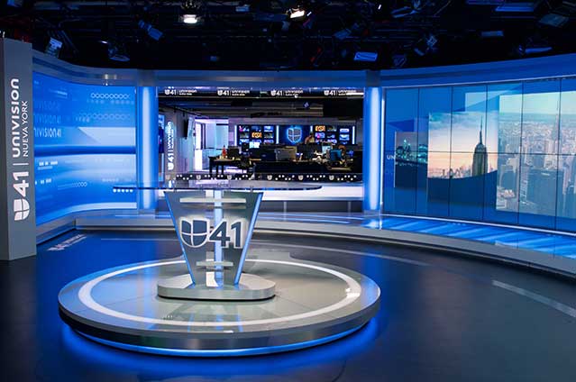 Modern Univision 41 news studio with a glass anchor desk, blue lighting accents, advanced video displays showing news graphics, and a newsroom visible in the background.