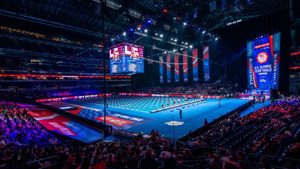 An indoor stadium filled with spectators features a brightly lit swimming pool and large video screens displaying “U.S. Olympic Team Trials,” with American flags and banners hanging above the pool.