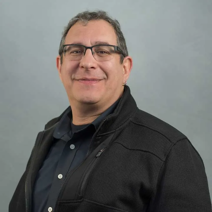 A man with short dark hair and glasses, wearing a black zip-up jacket over a dark shirt, smiles slightly while posing in front of a plain gray background with balanced lighting.